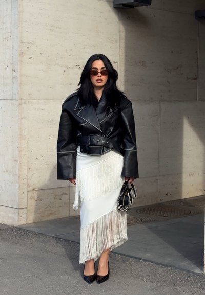 Mujer con chaqueta de cuero negra extragrande, falda blanca con flecos, tacones negros puntiagudos, gafas de sol y sosteniendo un pequeño bolso a rayas de cebra.