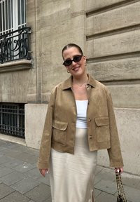 Vrouw in een beige jas, witte top en crèmekleurige rok, met een zwarte handtas, staande op de stoep van de stad voor een stenen gebouw.