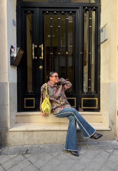 Mujer con gafas de sol y chaqueta estampada sentada en un bordillo de piedra frente a una puerta negra, sosteniendo un bolso amarillo, con una pierna cruzada sobre la otra.