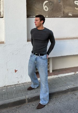 Hombre con camisa negra de manga larga y jeans azul claro está en la acera mirando hacia la izquierda, frente a una pared blanca con los números 5 y 6 descoloridos.