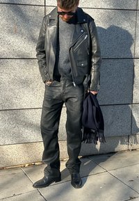 Fekete bőr motoros dzseki, szürke pulóver, fekete farmernadrág és fekete csizmák. Kézben tengerészkék sál. Láthatóak a texturált felületek, cipzárak és zsebek.