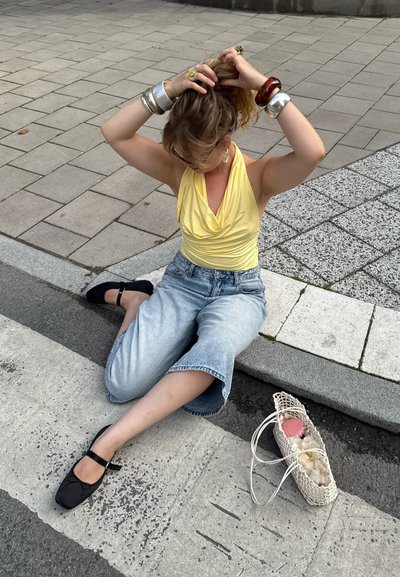 Top amarillo drapeado, culottes de mezclilla anchos de color azul claro, zapatos planos negros, accesorios de plata y madera, bolsa tejida blanca con un artículo rosa dentro.