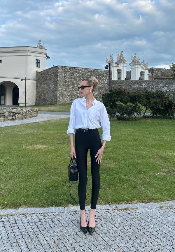 Camisa blanca de botones, leggings negros, bolso negro y tacones de punta. Fondo de hierba y piedra con características arquitectónicas. Cielo nublado.