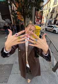 Casaco de couro castanho, suéter de malha e calças cinzentas. Mãos exibem vários anéis: dourados com pedras coloridas, designs redondos e quadrados. Unhas pintadas de vermelho.