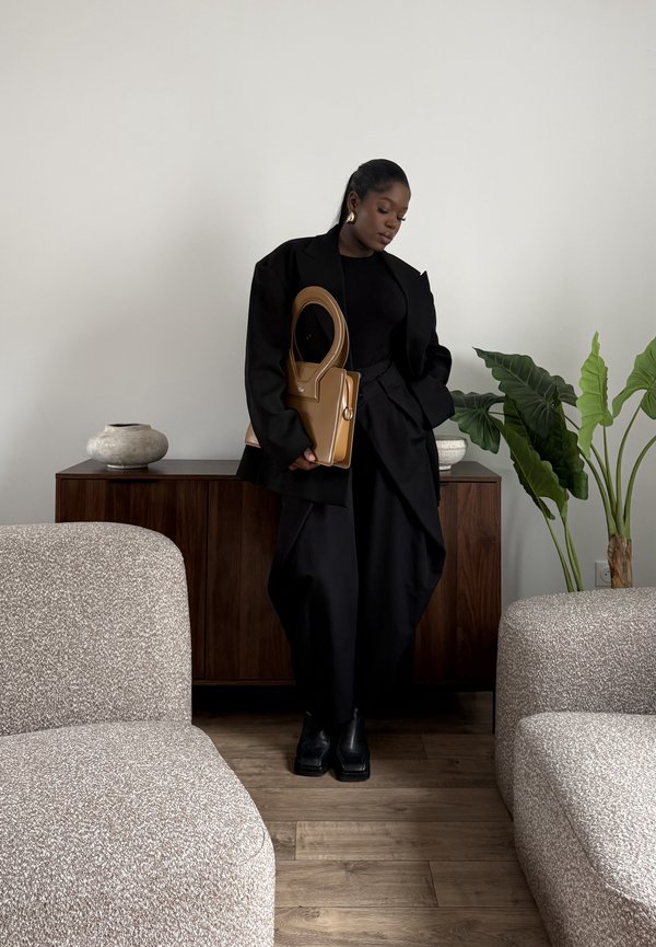 Donna con blazer nero oversize e pantaloni, che tiene una borsa marrone, in piedi tra due sedie beige dalla superficie strutturata in una stanza minimalista con piante.