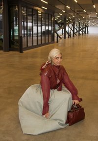 Femme portant un haut bordeaux, un foulard beige et une longue jupe, accroupie sur un grand sol intérieur en béton, tenant un sac à main bordeaux.