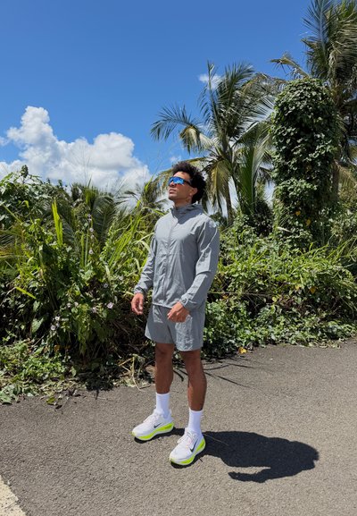 Hombre que lleva un conjunto deportivo gris y gafas de sol, de pie en la carretera cerca de plantas verdes y frondosas bajo un cielo azul brillante.