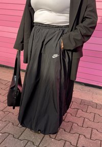 Saia preta Nike com cintura elástica, apresentando um bolso lateral e material leve e suave. Usada com uma camisola branca e um casaco preto.