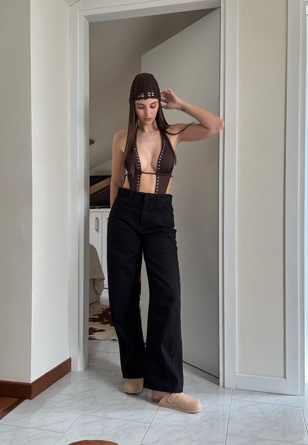 Femme portant un haut de bikini sombre, un pantalon noir taille haute à jambes larges, des chaussons beiges et un bonnet marron crocheté, debout dans un cadre de porte à l'intérieur.