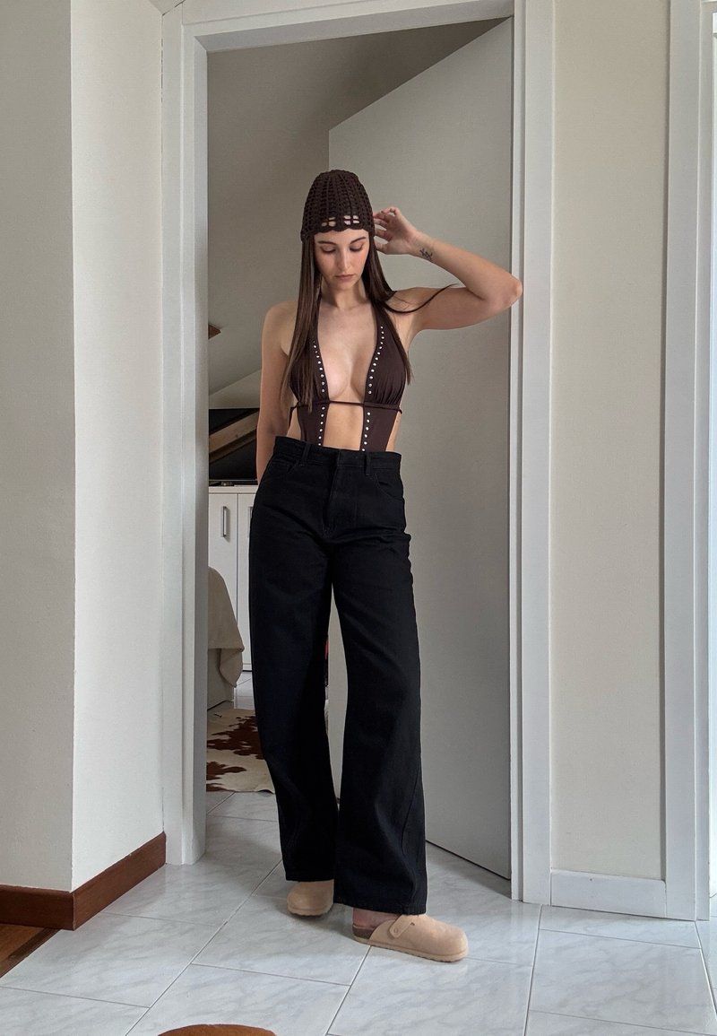 Femme portant un haut de bikini sombre, un pantalon noir taille haute à jambes larges, des chaussons beiges et un bonnet marron crocheté, debout dans un cadre de porte à l'intérieur.