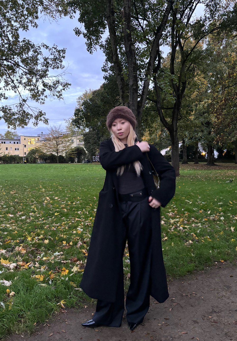 Čierny dvojradový kabát, tmavý top a široké nohavice. Hnedý kožušinový klobúk; špicaté čierne podpätky. Tráva a spadlé listy v pozadí.