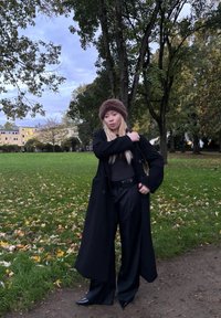 Čierny dvojradový kabát, tmavý top a široké nohavice. Hnedý kožušinový klobúk; špicaté čierne podpätky. Tráva a spadlé listy v pozadí.