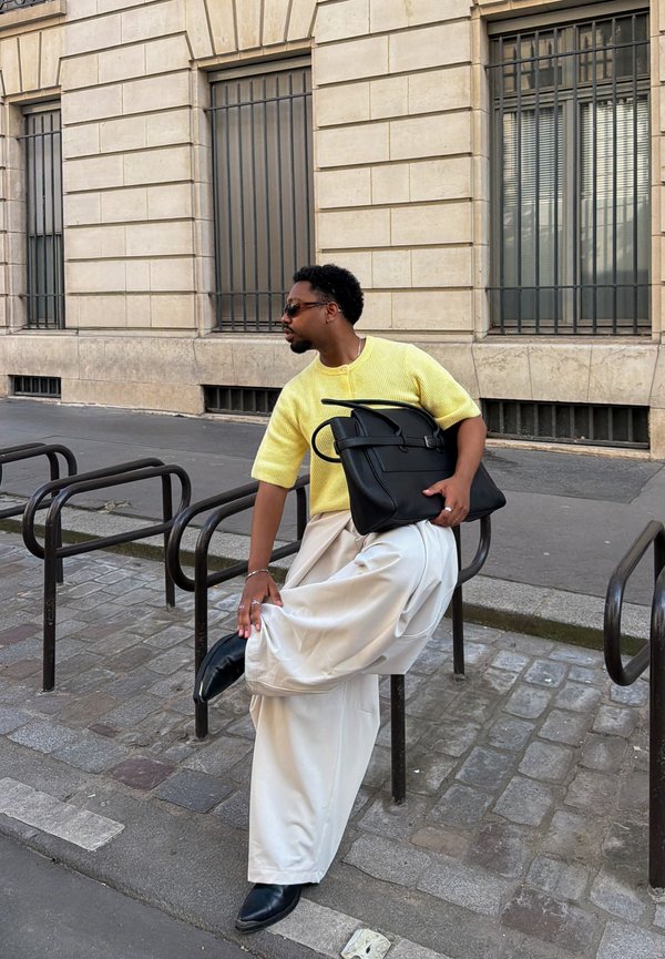 Homme en pull jaune, pantalon large blanc et chaussures noires ajuste son pied sur le porte-vélos sur le trottoir de la ville à côté d'un bâtiment en pierre.