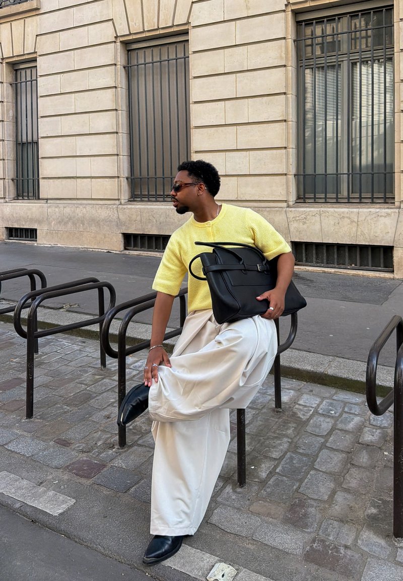 Homme en pull jaune, pantalon large blanc et chaussures noires ajuste son pied sur un support à vélos sur le trottoir en ville à côté d'un bâtiment en pierre.
