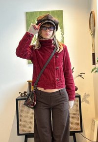 Femme portant une veste rouge, un chapeau à carreaux, des lunettes et un pantalon rayé se tient à l'intérieur près d'une armoire et d'une raquette de tennis vintage.