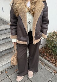 Jachetă maro căptușită cu blană, cu accente bej, cardigan verde deschis cu nasturi, pantaloni negri și ghete bej până la gleznă, ținând o geantă maro deschis.