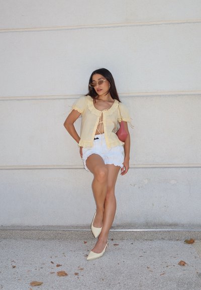 Blusa amarilla clara con bordes con volantes y detalle de lazo, combinada con pantalones cortos blancos con volantes y zapatos de slingback beige claro. Bolsa de mano rosa y gafas de sol.