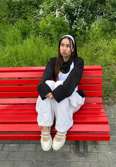 Joven mujer con cabello largo y oscuro y una cinta blanca en la cabeza sentada con las piernas cruzadas en un banco de parque rojo brillante, con vegetación detrás, vistiendo ropa negra y blanca.