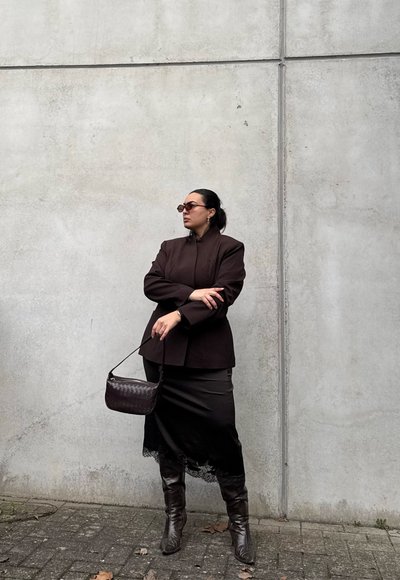 Mujer con blazer oscuro, falda negra, botas de cuero, gafas de sol, sosteniendo un bolso pequeño, de pie junto a una pared de hormigón.