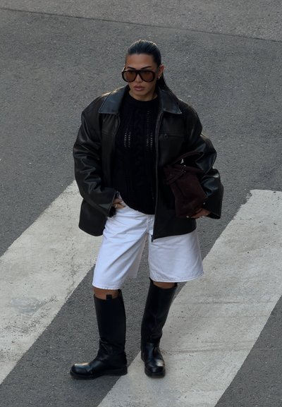 Chaqueta de cuero negra oversized, suéter negro de punto, pantalones cortos blancos y botas altas negras. Sosteniendo un bolso marrón, de pie en un paso de peatones.