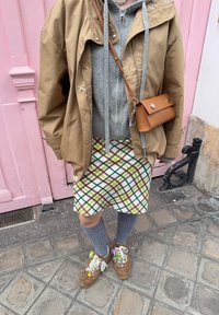 Kurtka w kolorze brązowym oversize, szary sweter, mini spódnica w kratę z zielonymi i brązowymi liniami, szare skarpetki do kolan, brązowe buty z kolorowymi akcentami.