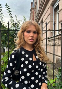 Sort cardigan med hvide prikker, parret med en matchende top. Har en rund halsudskæring og tekstureret stof. Baggrunden inkluderer grønt område og en bygning.