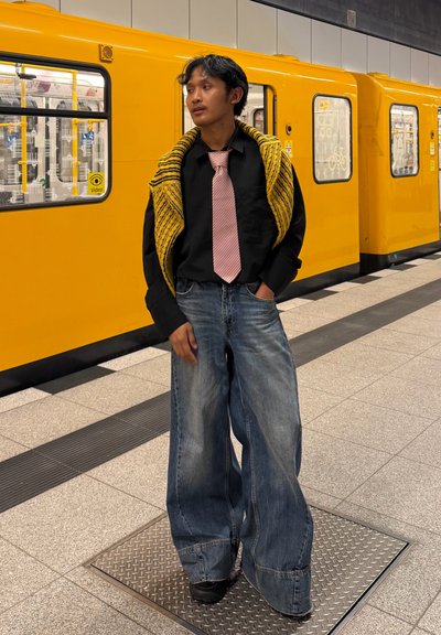 Persona con pantalones vaqueros de pierna ancha, camisa negra, corbata a rayas rosas y chal amarillo acanalado, de pie junto a un tren de metro amarillo.