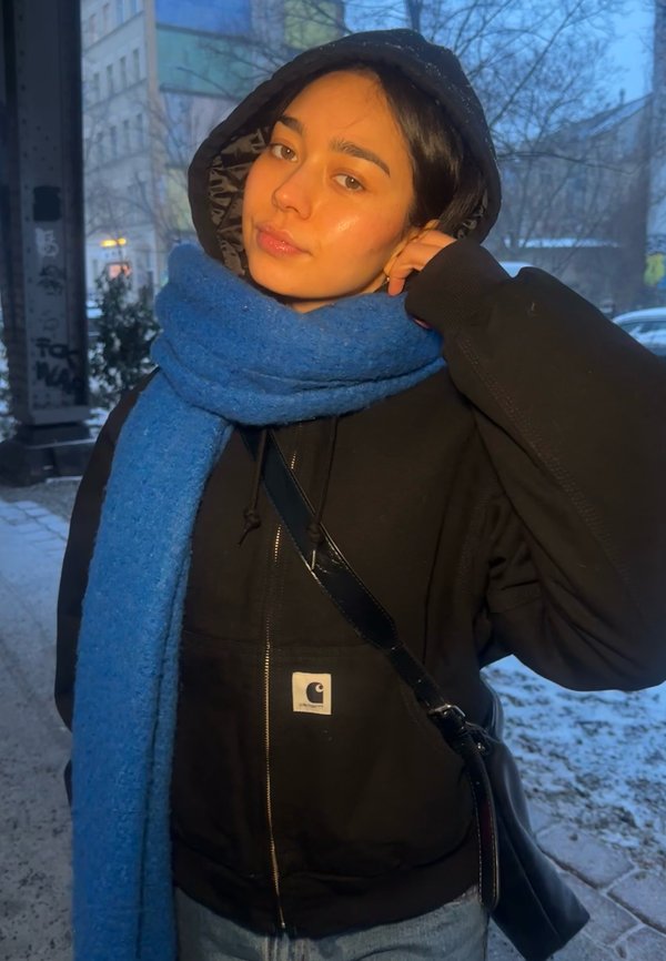 Junge Frau, die eine schwarze Kapuzenjacke, einen leuchtend blauen Schal und eine Umhängetasche trägt, steht draußen auf einer verschneiten städtischen Straße in der Dämmerung.