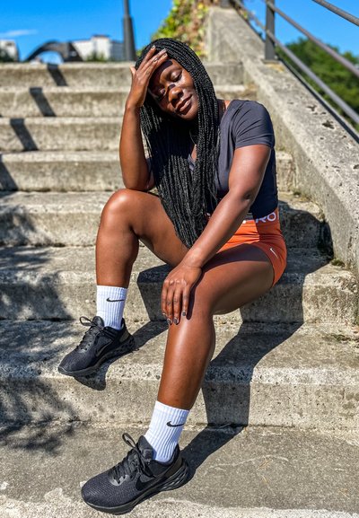 Zapatillas deportivas negras y calcetines blancos de Nike combinados con pantalones cortos naranjas y una camiseta negra. La persona está sentada en escalones de concreto bajo la luz del sol.