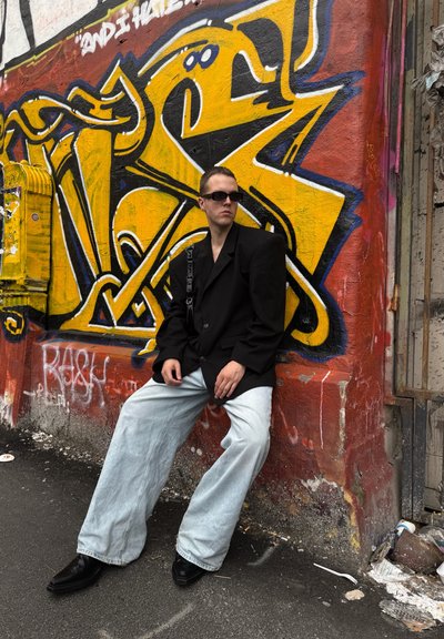 Blazer negro de gran tamaño, jeans de pierna ancha azul claro y botas de plataforma negras. Gafas de sol y un fondo de graffiti urbano añaden contraste.