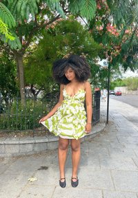 Vestido blanco con rayas verdes, textura con volantes y tirantes de espagueti ajustables. Combinado con zapatos planos de malla negra. Ambiente al aire libre con vegetación.