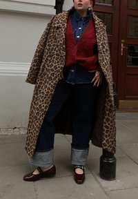 Manteau à imprimé léopard, gilet texturé rouge, chemise en denim, jeans foncés avec des ourlets clairs, et des chaussures Mary Jane bordeaux avec un détail de boucle.