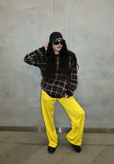 Mujer con gorra negra, gafas de sol, un suéter de cuadros negro con líneas amarillas y pantalones holgados de color amarillo brillante contra una pared gris.