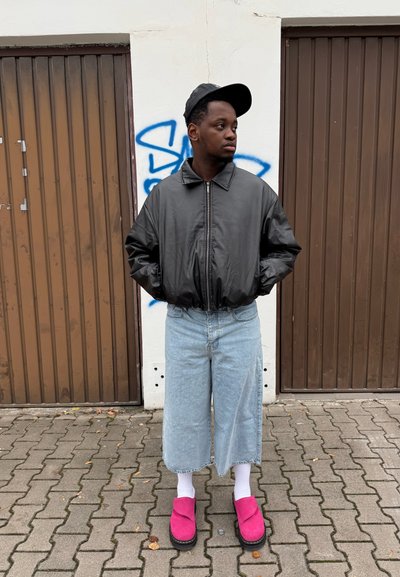 Chaqueta bomber negra, pantalones cortos de mezclilla anchos en azul claro, calcetines blancos, zapatillas rosas tipo slip-on. De pie sobre adoquines cerca de puertas de madera y graffiti.
