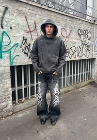 Grey zip-up hoodie with a hood, teamed with black baggy jeans that have large rips. Black shoes. The background features graffiti on a wall.