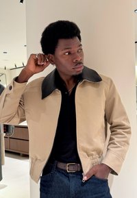 Beżowa kurtka z czarnym kołnierzem, z zapięciem na zamek i elastycznym paskiem w talii. Założona na czarną koszulę polo, w połączeniu z ciemnoniebieskimi dżinsami.