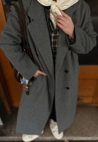 Szürke gyapjú keverék túlméretezett kabát nagy gombokkal, kockás ing fölött viselve. Világos színű sál van a nyak köré kötve, és egy barna táska lóg a vállon.