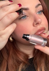 Producto cosmético en un recipiente transparente con un aplicador negro. Una persona aplica un producto brillante color rosa claro en el párpado.