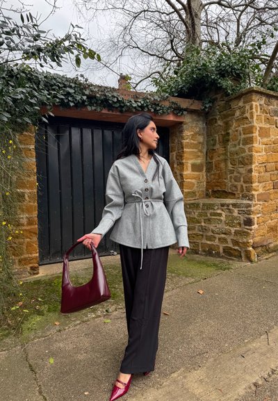 Chaqueta de color gris claro con cinturón y botones, combinada con pantalones de pierna ancha negros. Bolsa de mano de cuero burdeos y zapatos de punta a juego.