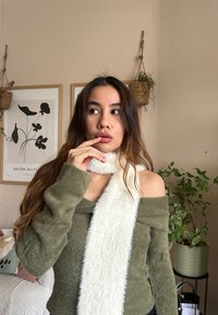 Grüner, flauschiger Off-Shoulder-Pullover kombiniert mit einem langen, flauschigen weißen Schal. Im Hintergrund sind hängende Pflanzen und gerahmte Kunstwerke zu sehen.