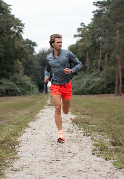 Camisa de running de manga larga gris y pantalones cortos naranja brillante, con un camino arenoso y un bosque verde de fondo. El calzado para correr es naranja.