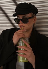 Fekete sapka lekerekített tetővel, nagy méretű fekete napszemüveg, hosszú körmök világos alappal és színes végekkel, egy átlátszó poharat tart zöld itallal.