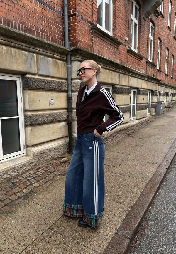 Frau mit blondem Dutt, schwarze Brille, dunkle Jacke, Jeans mit weitem Bein und karierten Bündchen, steht auf dem Kopfsteinpflaster neben einem Backsteingebäude.