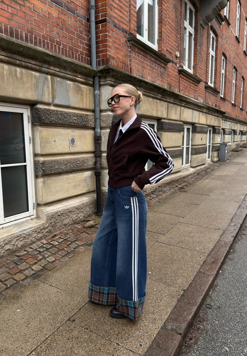 Femme aux cheveux blonds en chignon portant des lunettes noires, une veste sombre, un jean à jambes larges avec des revers à carreaux, debout sur un trottoir pavé près d'un bâtiment en briques.