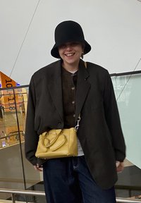 Svart bøttehatt, overdimensjonert svart blazer, brun skjorte med knapper, og denimbukser. Gul håndveske med to håndtak. Smilende uttrykk.