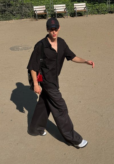 Camisa negra de gran tamaño, pantalones negros sueltos, zapatos blancos y gorra negra con un logo rojo. Lleva una bolsa de hombro con una etiqueta roja.