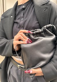 Melnas ādas striņķa soma ar savilkta augšu un sudraba detaļu akcentiem; turēta rokā ar garām, sarkaniem nagiem ar detaļām.