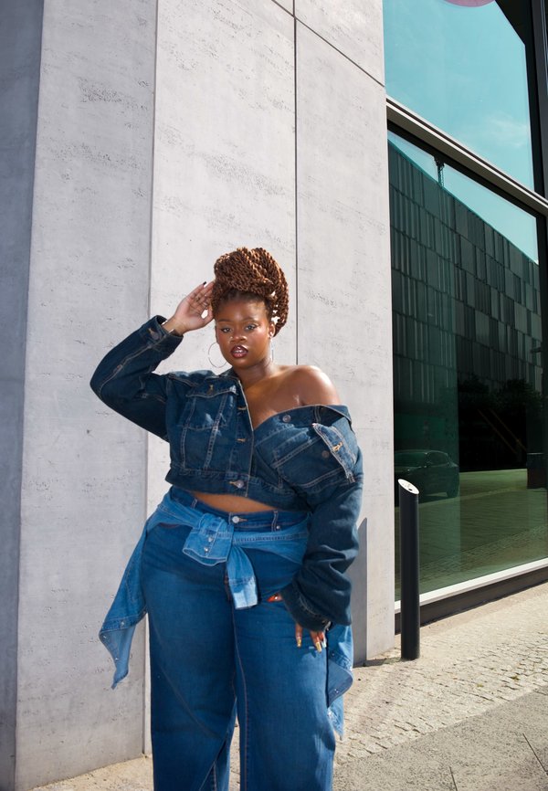 Femme aux cheveux tressés, vêtue d'une tenue en jean superposée, se tient contre un mur en béton près d'un bâtiment en verre, posant la main sur la tête.