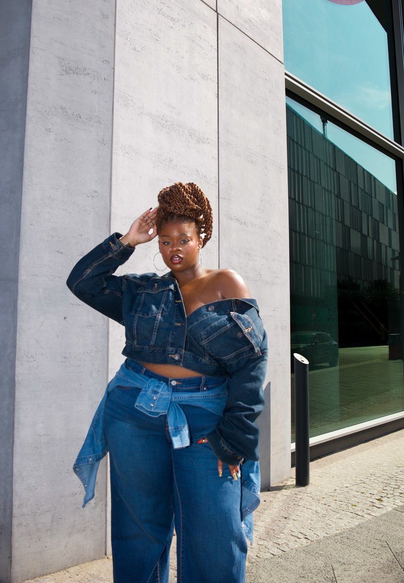 Femme aux cheveux tressés, vêtue d'une tenue en jean superposée, se tient contre un mur en béton près d'un bâtiment en verre, posant la main sur la tête.