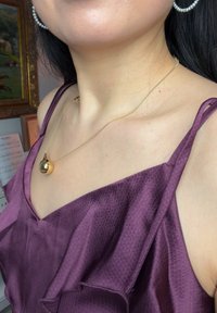 Γυναίκα που φοράει μωβ μπλούζα με βολάν, χρυσό κολιέ με στρογγυλό μενταγιόν και σκουλαρίκια κρίκους με πέρλες, με κορνιζαρισμένους πίνακες και παρτιτούρες στο φόντο.